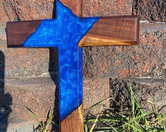 May include: A wooden cross with a blue resin inlay. The resin has a glittery finish. The cross is leaning against a brick wall.
