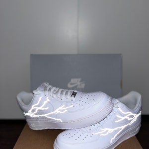 May include: White leather sneakers with reflective lightning bolt designs on the sides. The shoes are displayed on top of a brown cardboard box, with a gray Nike box in the background. The sneakers have white laces and soles.
