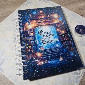 Op de afbeelding: Een blauw en zwart spiraalgebonden notitieboek met een glinsterende omslag met een boekenkast met een deuropening waarop "Er was eens" staat.