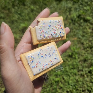 Puede incluir: Dos objetos rectangulares con forma de pastel con una corteza dorada y una capa superior de glaseado blanco. El glaseado está decorado con pequeñas chispas multicolores. Un pastel presenta una marca de mordisco visible en la esquina superior.