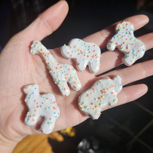 Könnte beinhalten: Fünf tierförmige Kekse mit weißer Glasur und bunten Streuseln. Die Kekse sind in Form einer Giraffe, eines Elefanten, eines Hundes, eines Nilpferds und eines Bären gehalten. Die Kekse werden in einer Hand gehalten.