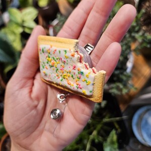 May include: A rectangular pastry-shaped badge reel featuring a golden-brown crust, white icing, and colorful sprinkles. The pastry has a visible bite mark on the top right corner, revealing a red filling. A retractable cord and metal clip are attached.