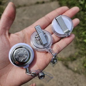 May include: Three round badge reels with white plastic housings and clear plastic straps. One reel shows a silver metal back with the word MAGNETIC embossed. The other two reels feature silver metal belt clips on their clear plastic backs.