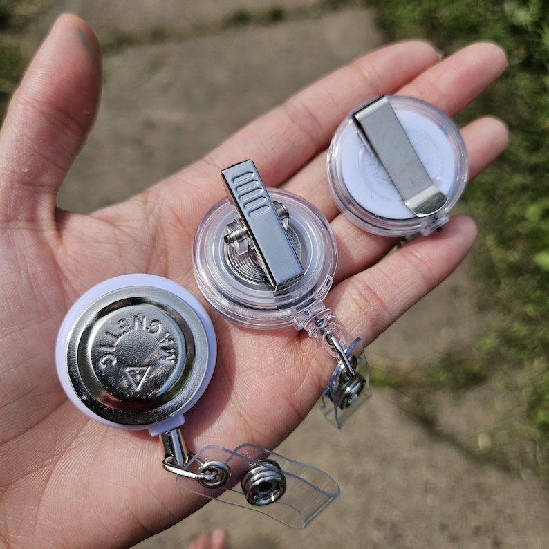 May include: Three round badge reels with white plastic housings and clear plastic straps. One reel shows a silver metal back with the word MAGNETIC embossed. The other two reels feature silver metal belt clips on their clear plastic backs.