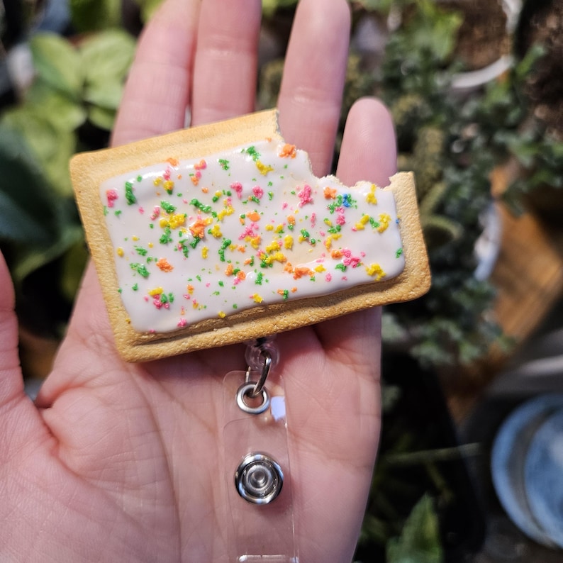 May include: A rectangular pastry-shaped badge reel featuring a golden-brown crust, white icing, and colorful sprinkles. The pastry has a visible bite mark on the top right corner and is attached to a clear plastic strap with a metal snap button.