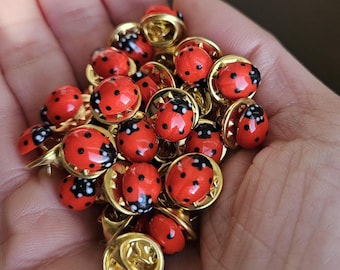 May include: A collection of ladybug-shaped lapel pins. Each pin features a red body with black spots and a gold-colored metal base. The pins are small and detailed, with a glossy finish.