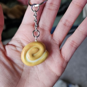 Può includere: Un portachiavi con un ciondolo a forma di spirale. Il ciondolo è giallo con una striscia bianca e una finitura lucida. Il portachiavi ha una catena e un anello in metallo argentato.