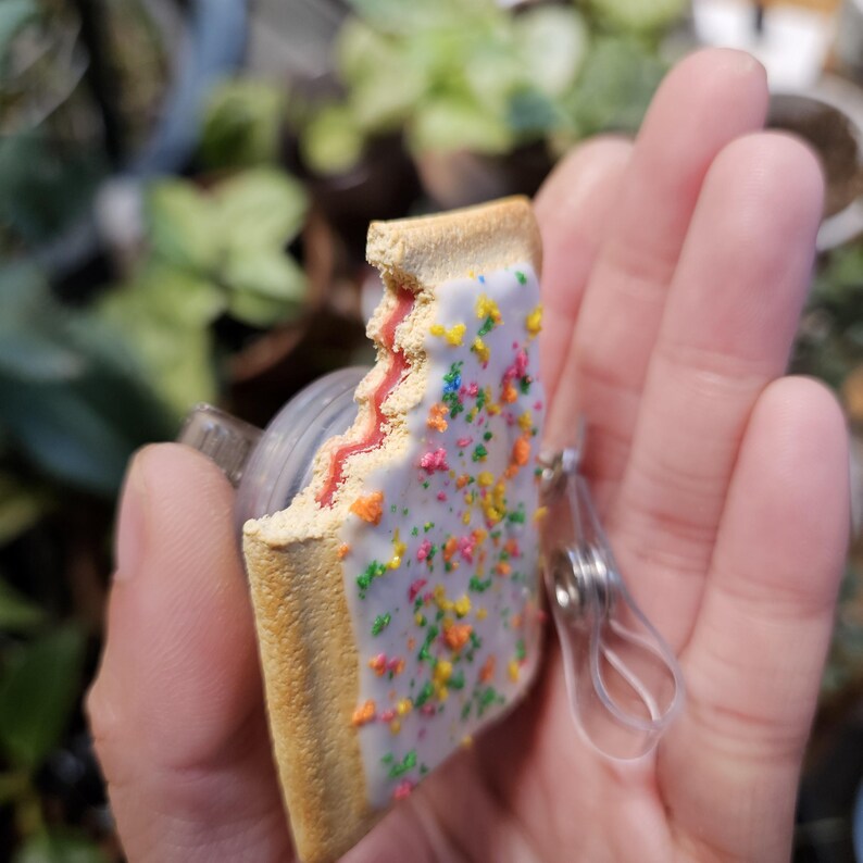 May include: A rectangular pastry-shaped badge reel featuring a light brown crust, a white frosted top with colorful sprinkles, and a visible red fruit filling layer. The item includes a clear plastic strap and a circular metal clip mechanism.