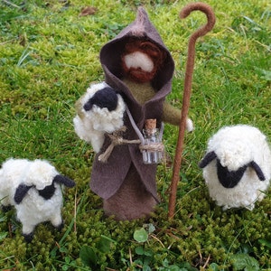 May include: A felted shepherd figure in a brown hooded cloak, holding a staff and a bag with a sheep. Two white sheep with black faces are nearby. A small glass bottle hangs from the shepherd's neck.
