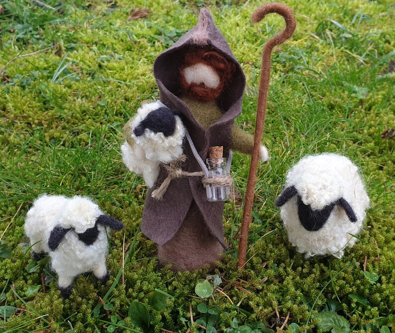 May include: A felted shepherd figure in a brown hooded cloak, holding a staff and a bag with a sheep. Two white sheep with black faces are nearby. A small glass bottle hangs from the shepherd's neck.