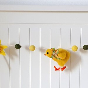 May include: A felt garland with a spring theme. It features a chick, carrots, a daffodil, and tulips. The garland is strung with yellow, orange, and green pom-poms.