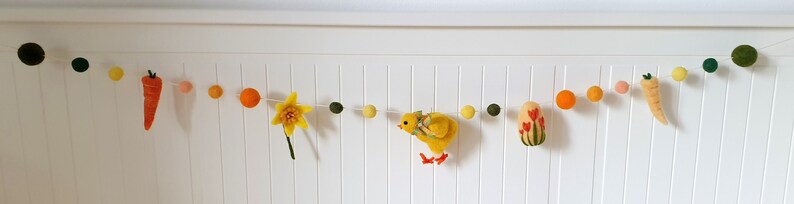 May include: A felt garland with a spring theme. It features a chick, carrots, a daffodil, and tulips. The garland is strung with yellow, orange, and green pom-poms.