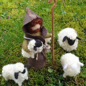 May include: A felted shepherd figure with a brown hooded cloak, holding a staff and a small lamb. Three white sheep with black markings are nearby, all set on a bed of green moss.