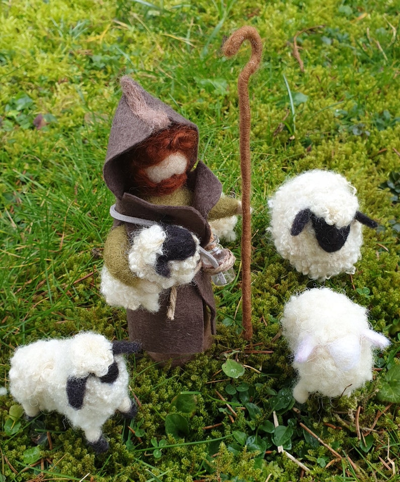 May include: A felted shepherd figure with a brown hooded cloak, holding a staff and a small lamb. Three white sheep with black markings are nearby, all set on a bed of green moss.