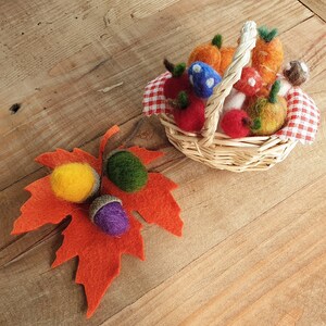 May include: A small wicker basket filled with felt fruit and vegetables, including red apples, orange pumpkins, and mushrooms. The basket is decorated with a red and white checked fabric. A felt maple leaf with three acorns is on the wooden surface.