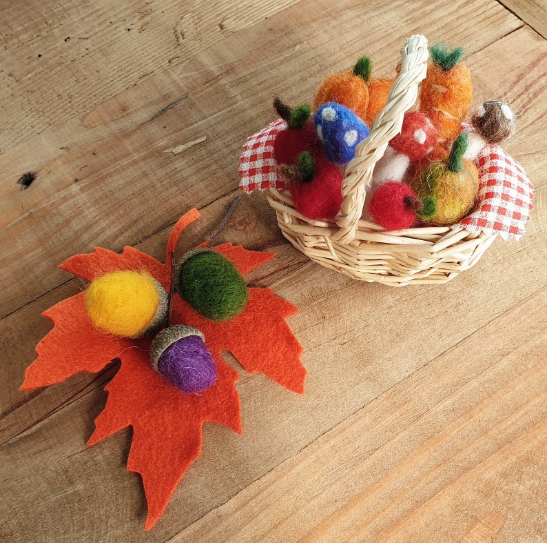 May include: A small wicker basket filled with felt fruit and vegetables, including red apples, orange pumpkins, and mushrooms. The basket is decorated with a red and white checked fabric. A felt maple leaf with three acorns is on the wooden surface.