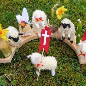 May include: A wooden arch holds felted wool figures of lambs, a daffodil, and an Easter egg. The lambs have red bows and some hold red flags with white crosses. A green candle with white flowers is also present.