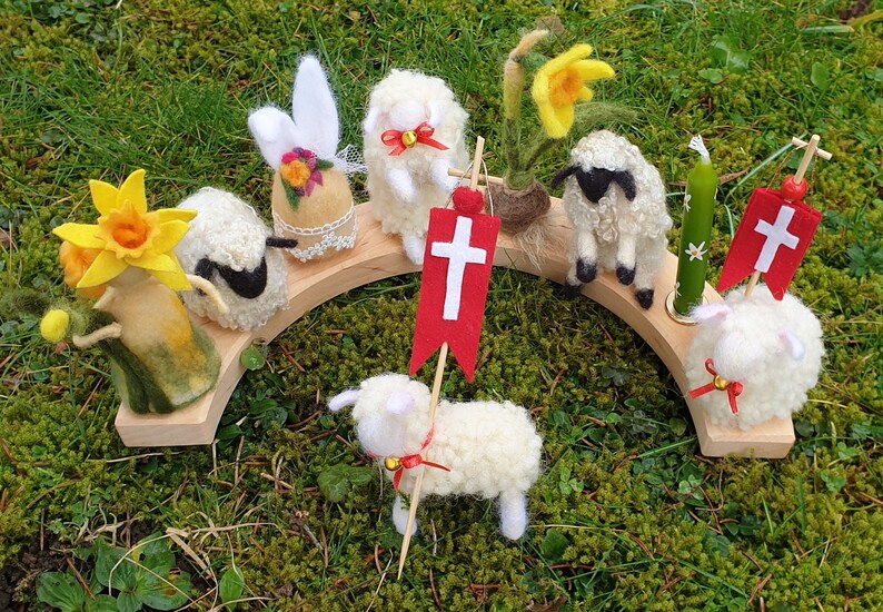 May include: A wooden arch holds felted wool figures of lambs, a daffodil, and an Easter egg. The lambs have red bows and some hold red flags with white crosses. A green candle with white flowers is also present.
