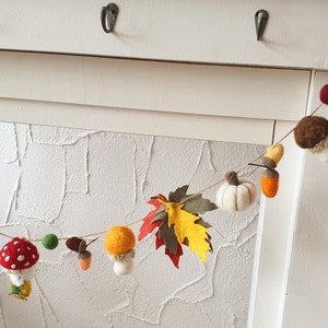 May include: A felt garland with autumnal themes, featuring mushrooms, leaves, acorns, and pumpkins in shades of red, orange, yellow, brown, and green.