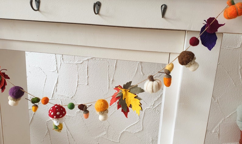 May include: A felt garland with autumnal themes, featuring mushrooms, leaves, acorns, and pumpkins in shades of red, orange, yellow, brown, and green.