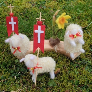 May include: A whimsical Easter scene featuring three fluffy, white wool sheep figurines with red ribbon accents and small gold bells. Two sheep hold red banners with white crosses. A yellow daffodil and a wooden platform complete the pastoral display.