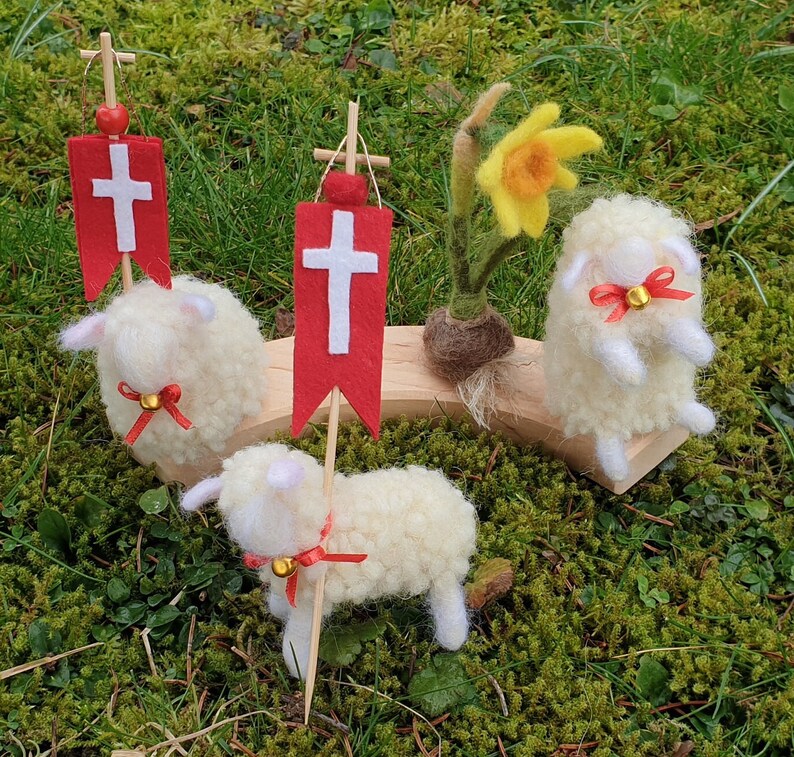 May include: A whimsical Easter scene featuring three fluffy, white wool sheep figurines with red ribbon accents and small gold bells. Two sheep hold red banners with white crosses. A yellow daffodil and a wooden platform complete the pastoral display.