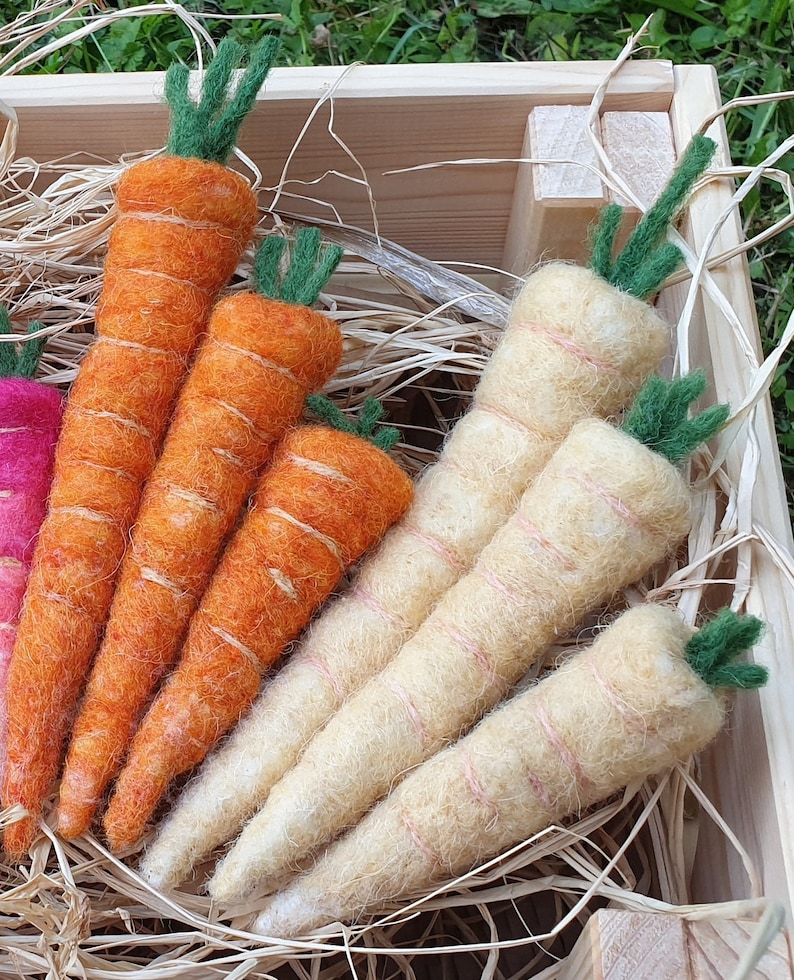 May include: A close-up of a wooden crate filled with felt carrots and parsnips. The carrots are orange and the parsnips are a pale yellow. The vegetables are arranged in a visually appealing way.