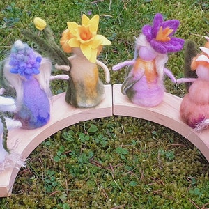 May include: A whimsical display of felted flower figures in various colors, including white, yellow, purple, and pink. They are arranged on a wooden arch, creating a charming, handcrafted scene. The figures have delicate features and are set against a backdrop of green foliage.