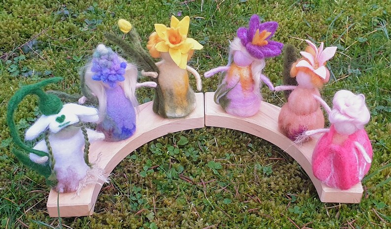 May include: A whimsical display of felted flower figures in various colors, including white, yellow, purple, and pink. They are arranged on a wooden arch, creating a charming, handcrafted scene. The figures have delicate features and are set against a backdrop of green foliage.