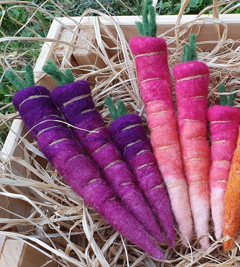 May include: A set of hand-felted carrots in a variety of colors, including purple, pink, and orange. The carrots are arranged in a wooden crate filled with straw.