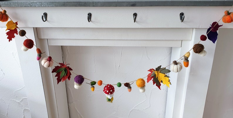 May include: A felt garland with autumnal themes, featuring pumpkins, leaves, acorns, mushrooms, and a red and white mushroom with a white stem. The garland is strung on a white background.