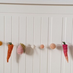 May include: A felt garland with a string of colorful pom-poms and felt carrots and eggplant. The garland is hung on a white wall.