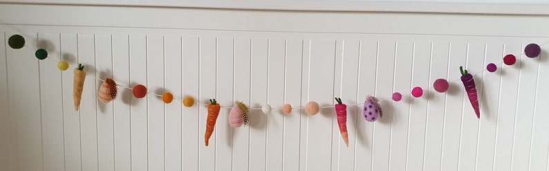 May include: A felt garland with a string of colorful pom-poms and felt carrots and eggplant. The garland is hung on a white wall.