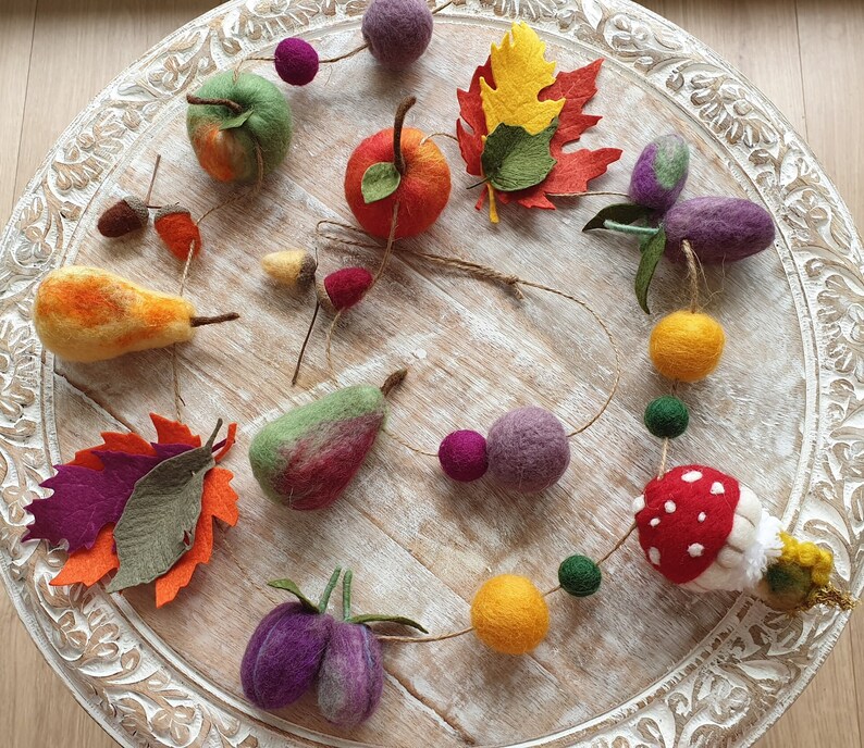 May include: A felt garland with an autumn theme. It features felt leaves, fruit, and other shapes in various colours, including red, orange, yellow, green, purple, and brown.