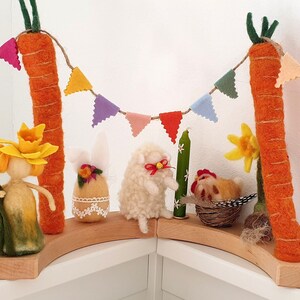 May include: A felt Easter scene with a bunny, lamb, chicken, and a girl with a daffodil crown. The scene is decorated with a string of colorful felt flags and two orange felt carrots.