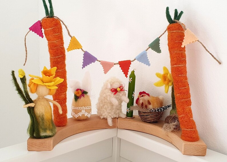 May include: A felt Easter scene with a bunny, lamb, chicken, and a girl with a daffodil crown. The scene is decorated with a string of colorful felt flags and two orange felt carrots.