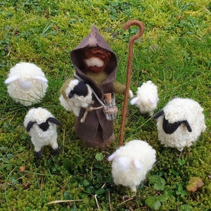 May include: A felted shepherd figure in a brown hooded cloak, holding a staff and a small glass bottle, surrounded by six white and black sheep figurines. The scene is set on a bed of green moss.