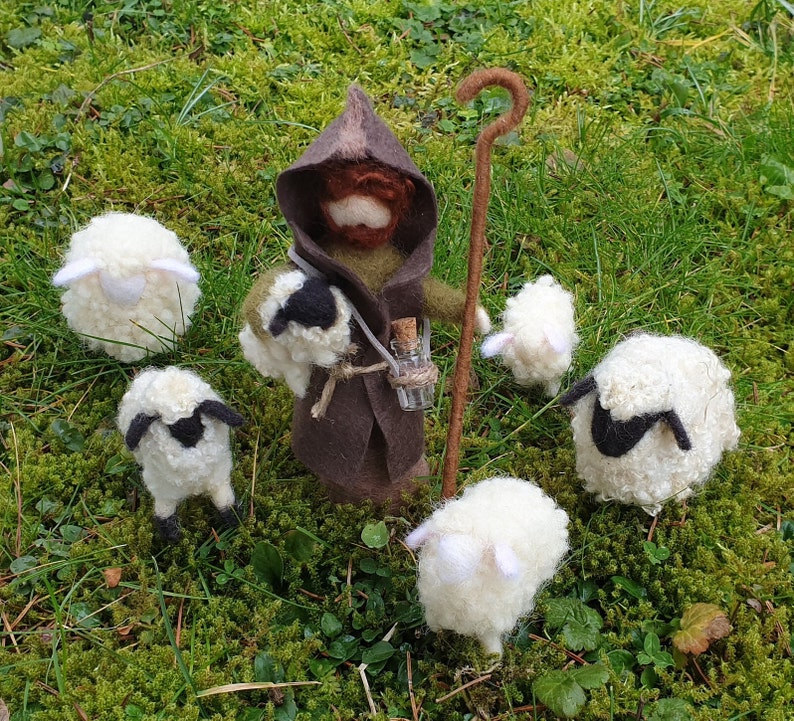 May include: A felted shepherd figure in a brown hooded cloak, holding a staff and a small glass bottle, surrounded by six white and black sheep figurines. The scene is set on a bed of green moss.