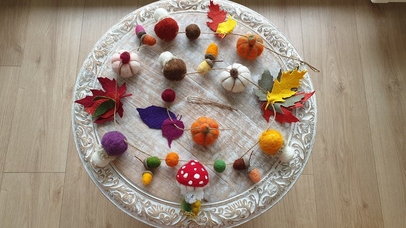May include: A felt garland featuring autumnal decorations, such as pumpkins, leaves, acorns, and a mushroom. The garland is strung on white twine and is displayed on a white wooden table.