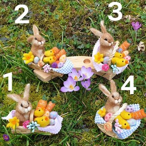May include: Four felt Easter bunnies, each holding a basket filled with colorful eggs and chicks. The bunnies are sitting on a wooden platform with a blue and white checkered tablecloth. The background is a green grassy area with purple flowers.