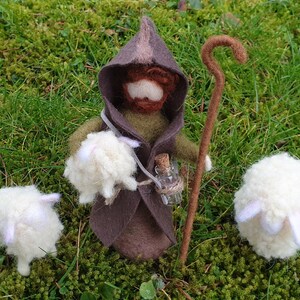 May include: A handcrafted felt shepherd doll with a brown hooded cloak, holding a staff and a small bottle. Two white sheep are positioned on either side. The scene is set on a bed of green moss and grass.