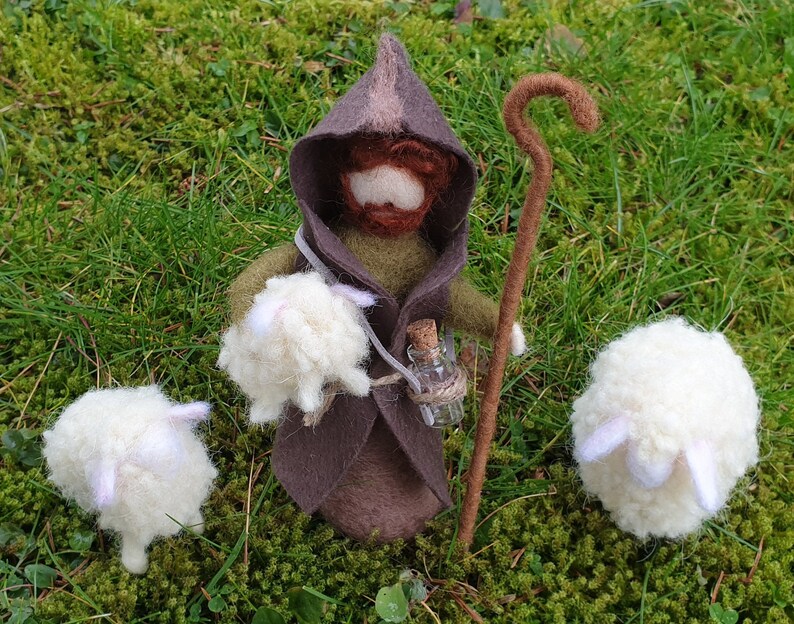 May include: A handcrafted felt shepherd doll with a brown hooded cloak, holding a staff and a small bottle. Two white sheep are positioned on either side. The scene is set on a bed of green moss and grass.