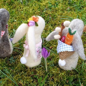 May include: Five handmade felt Easter bunnies with different colored fur and floral headbands. Each bunny is holding a basket filled with spring-themed items like carrots, flowers, and eggs.