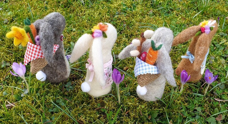 May include: Five handmade felt Easter bunnies with different colored fur and floral headbands. Each bunny is holding a basket filled with spring-themed items like carrots, flowers, and eggs.