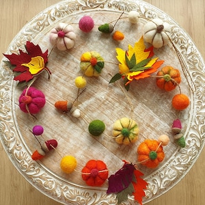 May include: A felt garland featuring autumnal decorations, such as pumpkins, leaves, acorns, and balls in a range of colours. The garland is strung on white twine and is displayed on a white wooden surface.