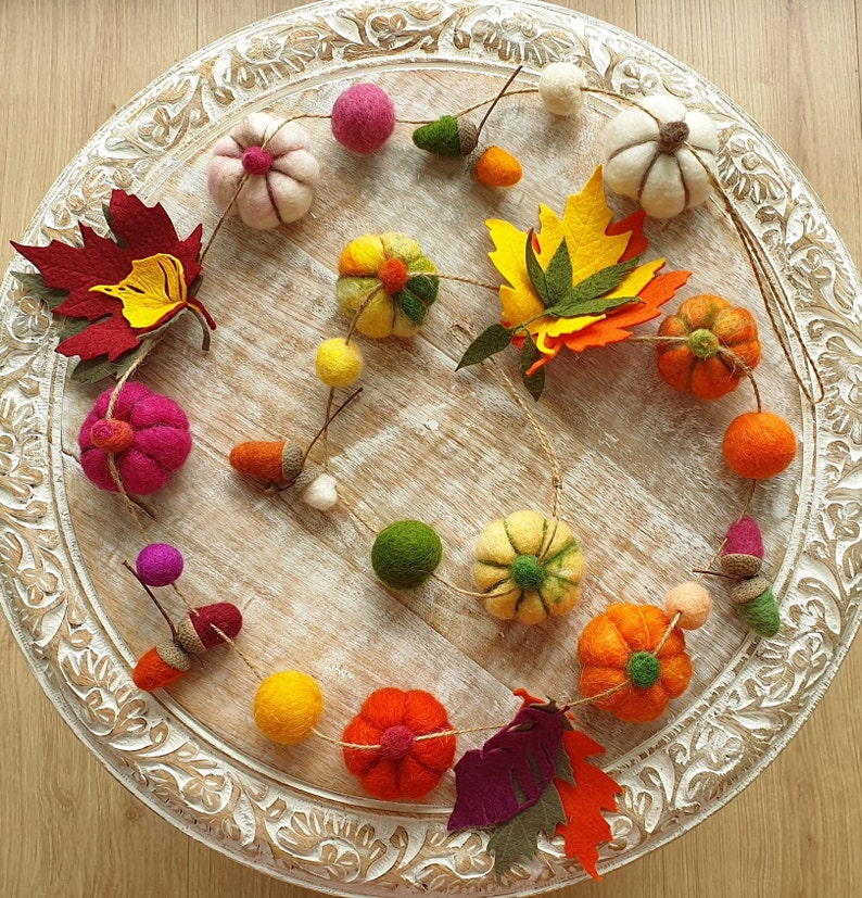 May include: A felt garland featuring autumnal decorations, such as pumpkins, leaves, acorns, and balls in a range of colours. The garland is strung on white twine and is displayed on a white wooden surface.