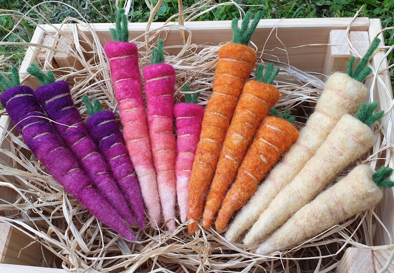 May include: A collection of hand-felted carrots in various colours, including purple, pink, orange, and cream. The carrots are arranged in a wooden crate with straw.
