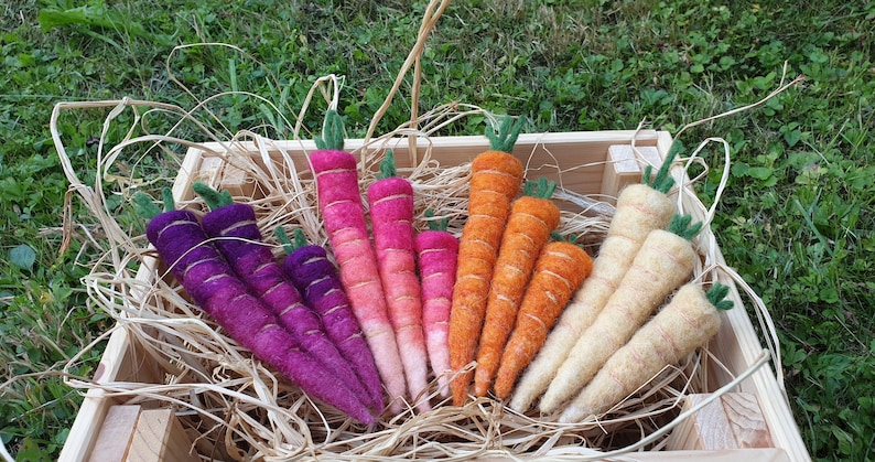 May include: A collection of hand-felted carrots in a wooden crate. The carrots are arranged in rows by color: purple, pink, orange, and cream.