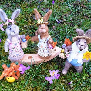 May include: Three felt bunny rabbits dressed in Easter outfits with baskets of carrots and eggs. The bunnies are sitting on a wooden swing set in a field of green grass and purple flowers.