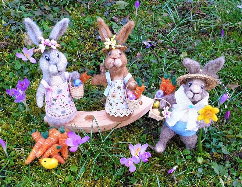 May include: Three felt bunny rabbits dressed in Easter outfits with baskets of carrots and eggs. The bunnies are sitting on a wooden swing set in a field of green grass and purple flowers.